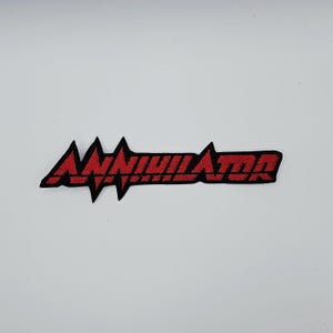 May include: Embroidered patch with the word "ANNIHILATOR" in red, stylized text. The text is outlined in black and set against a white background. The patch is rectangular and likely designed to be ironed or sewn onto fabric.
