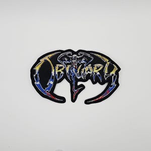 May include: Black embroidered patch with the word "OBITUARY" in a stylized font. The patch features a central graphic of a skeletal figure with wings, and is outlined in gold, blue, and red.
