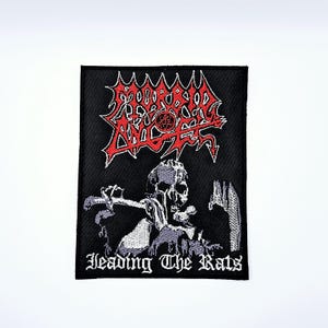 Op de afbeelding: Zwart geborduurde patch met een rood en wit logo en een skeletfiguur. De tekst "HEADING THE RATS" wordt in witte letters onder de afbeelding weergegeven. De patch is rechthoekig met een zwarte rand.