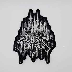 以下が含まれることがあります： ゴシック体の「Dark Fortress」の文字と城のイラストが施された、白と黒の刺繍パッチ。パッチは不規則な形状をしており、衣類やアクセサリー用としてデザインされている可能性があります。