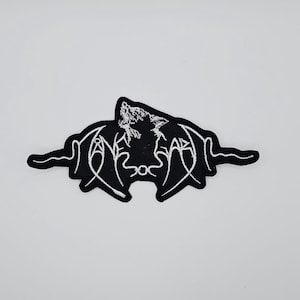 Könnte beinhalten: Schwarz-weißer, gestickter Aufnäher mit einem heulenden Wolf, stilisierten Flügeln und Text. Der Aufnäher hat einen schwarzen Hintergrund mit weißen Nähten, was einen auffälligen Kontrast erzeugt. Das Design ist symmetrisch und detailliert.