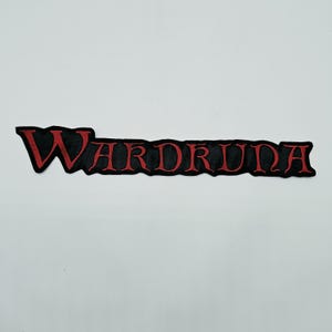 May include: A long, horizontal embroidered patch featuring the word WARDRUNA in stylised, bold red lettering with a black outline. The letters are connected in a continuous, jagged-edged design, creating a distinct, sharp-angled appearance.