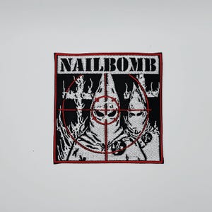 Puede incluir: Parche cuadrado con la palabra "NAILBOMB" en letras blancas sobre fondo negro. El parche presenta un gráfico de dos figuras encapuchadas dentro de una retícula de objetivo roja. Las figuras son blancas y el fondo es negro.