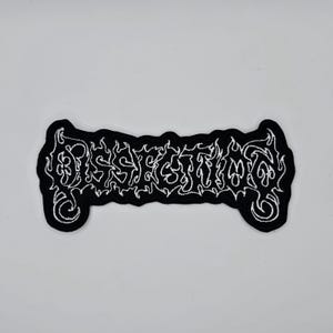 May include: Black embroidered patch with the word "Dissection" in white, stylized, gothic lettering. The patch has a unique shape with rounded ends and a black border. The design is intricate and detailed.