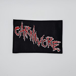 May include: Black rectangular patch with the word "CARNIVORE" embroidered in red with white outlining. The text has a jagged, distressed font style. The patch is likely for clothing or accessories.