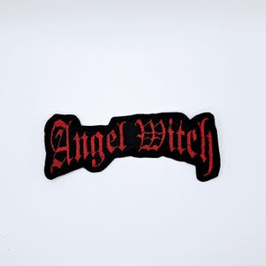 May include: Black embroidered patch featuring the words "Angel Witch" in red gothic script. The patch is designed with a distinctive shape and is likely intended for application to garments or accessories.