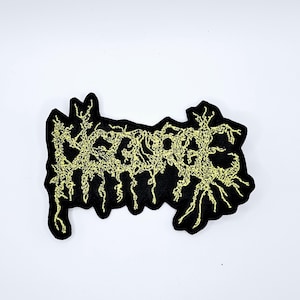 May include: Black embroidered patch with the word "DEGRADE" in a stylized, yellow, gothic font. The patch has a jagged, irregular border and a detailed, textured design.