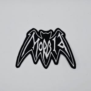 May include: A black and white embroidered patch featuring a bat design with the word "MORBID" in stylized white lettering. The patch has a black border and is likely designed to be ironed or sewn onto clothing or accessories.