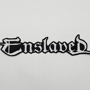 May include: A black and white embroidered patch with the word "Enslaved" in a gothic font. The patch has a black background with white stitching outlining the letters. The patch is rectangular with a pointed end.