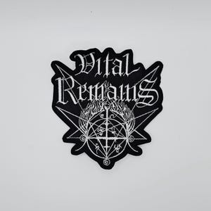 Puede incluir: Parche bordado en blanco y negro con las palabras "Vital Remains" en letras góticas. El parche incluye un pentagrama y otros símbolos ocultos, con un diseño de llama en el centro. Probablemente merchandising de una banda o artículo relacionado.