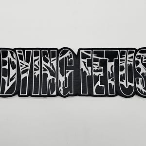 Puede incluir: Parche bordado en blanco y negro con el texto "DYING FETUS". El texto está en una fuente estilizada con detalles blancos dentro de las letras negras. El parche tiene un borde negro.