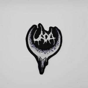 以下が含まれることがあります： 白とグレーのデザインの黒いパッチ。パッチには、三日月型の中に様式化されたフォントで「UADA」という単語が使用されています。パッチの下部には滴るような効果があります。パッチはおそらく衣類やアクセサリー用です。