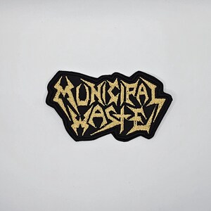 May include: Black embroidered patch with the words "MUNICIPAL WASTE" in gold thread. The patch has a jagged, irregular border. The text is in a stylized, heavy metal font.