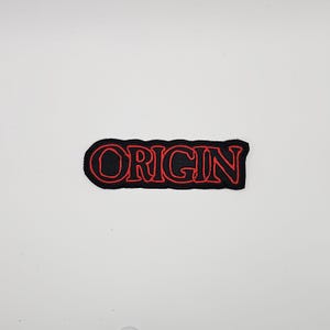 Puede incluir: Un parche bordado negro con la palabra "ORIGIN" en letras rojas. El parche tiene un borde negro y está sobre un fondo blanco. Las letras están en una fuente audaz y estilizada.