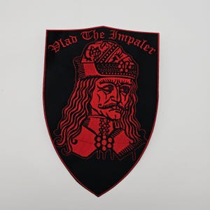 Può includere: Toppa nera a forma di scudo con un ritratto ricamato in rosso di Vlad l'Impalatore. Il testo "Vlad The Impaler" è arcuato in alto in rosso. La toppa presenta una dettagliata raffigurazione del volto, della corona e dell'abbigliamento di Vlad.