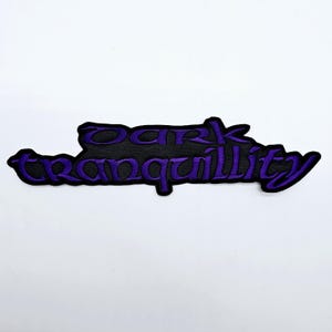 Könnte beinhalten: Ein schwarzer, gestickter Aufnäher mit dem Schriftzug "DARK tranquility" in Lila. Der Aufnäher hat einen schwarzen Hintergrund und einen lila Rand, vor weißem Hintergrund.