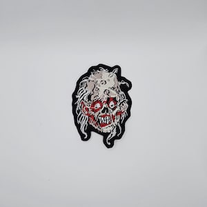 Könnte beinhalten: Gestickter Aufnäher mit einem Zombiegesicht mit roten Augen und Mund, weißen Haaren und einem schwarzen Rand. Der Aufnäher hat ein detailliertes Design, ideal zum Aufnähen auf Kleidung oder Accessoires.