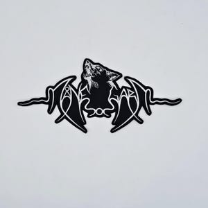 May include: Black and white patch featuring a howling wolf silhouette. The design includes stylized text and wing-like shapes, creating a gothic or fantasy aesthetic. The patch is likely for clothing or accessories.