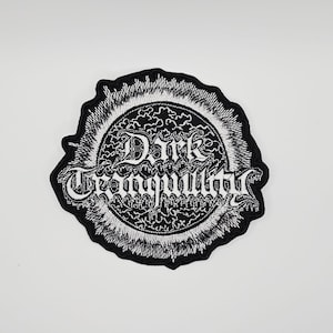 Könnte beinhalten: Gestickter Aufnäher mit dem Schriftzug "Dark Tranquillity" in weiß. Der Aufnäher hat einen schwarzen Rand und ein kreisförmiges Design mit einem detaillierten, wirbelnden Muster in Weiß. Geeignet für Kleidung oder Accessoires.