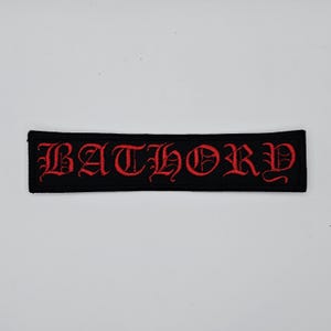 May include: Black rectangular patch with the word "BATHORY" embroidered in red gothic lettering. The patch is a rectangle with a black border and the red text is centered.