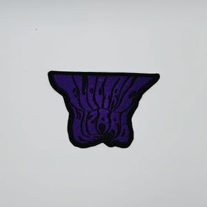 Puede incluir: Un parche bordado morado con un borde negro. El parche presenta texto estilizado en un diseño arremolinado. El parche está sobre un fondo blanco.