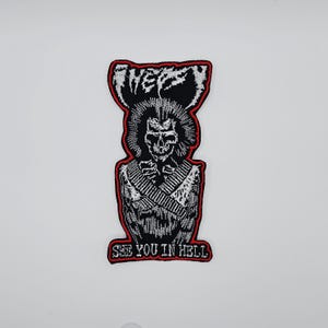 Può includere: Toppa ricamata con un disegno a teschio, capelli lunghi e un gilet in stile militare. La toppa recita "WEP" sopra il teschio e "SEE YOU IN HELL" sotto, in lettere bianche con un bordo rosso. Il design è principalmente in bianco e nero.