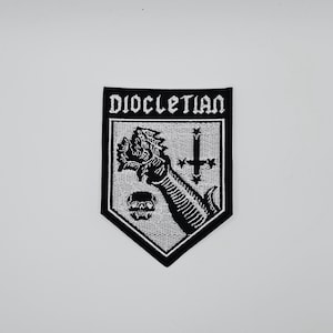 Puede incluir: Parche bordado en blanco y negro con la palabra "DIOCLETIAN" en la parte superior. El parche presenta un brazo esquelético sosteniendo una cabeza de dragón, una calavera y una cruz invertida.