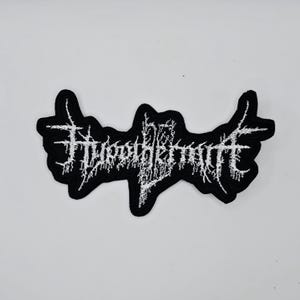 May include: Black and white embroidered patch with the text "FURTHERMITE" in a gothic font. The patch has a unique, irregular shape with pointed edges. The text is in a stylized, jagged font.