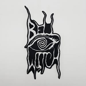 May include: A black and white embroidered patch with the words "BE WITCH" and a stylized eye. The design has a dripping, gothic aesthetic, suitable for clothing or accessories.