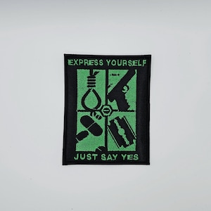 Pode incluir: Patch preto e verde com as frases "EXPRESS YOURSELF" e "JUST SAY YES". O design do patch inclui uma forca, uma arma, pílulas e uma lâmina de barbear dispostas em uma grade.