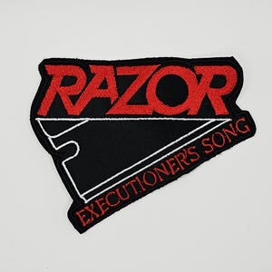 May include: A black embroidered patch featuring the word RAZOR in bold red lettering above a white outline of a razor blade shape. The text EXECUTIONER'S SONG is embroidered in red along the bottom edge of the patch against a black background.