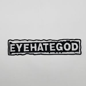 Puede incluir: Un parche bordado en blanco y negro con el texto "EYEHATEGOD". El texto blanco está en letras de bloque sobre un fondo rectangular negro. El parche tiene un borde blanco desgastado.