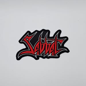 Puede incluir: Parche bordado con la palabra "SAVAGE" en letras rojas estilizadas. El texto está delineado en blanco y sobre un fondo negro. El parche tiene una forma irregular y dentada.