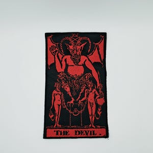 Puede incluir: Un parche rectangular bordado con un diseño en rojo y negro de la carta del tarot El Diablo. El gráfico muestra una figura con cuernos y alas, un pentagrama sobre la cabeza y dos figuras encadenadas. El texto THE DEVIL aparece en la parte inferior.