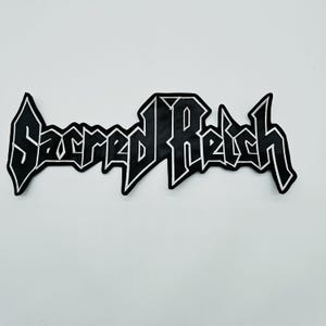 以下が含まれることがあります： 白い縁取りが施された、角張ったスタイリッシュなフォントで「Sacred Reich」という文字が刺繍された黒いワッペン。文字は太くシャープで、連続したデザインで繋がっており、ロゴのような独特の外観を作り出しています。