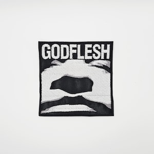 May include: A square, black patch with the white text "GODFLESH" at the top. Below the text is a white and black abstract design. The patch is likely for clothing or accessories.