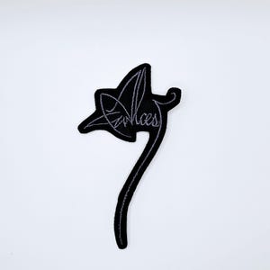 May include: Black embroidered patch in the shape of a leaf with the word "Voices" in cursive script. The patch has a black background with gray stitching and a long stem.