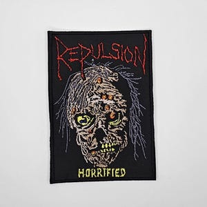 Może przedstawiać: Czarna, haftowana naszywka z motywem czaszki zombie, żółtymi oczami i napisem "Repulsion" w kolorze czerwonym. Słowo "Horrified" jest wyhaftowane na żółto pod czaszką.