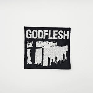 May include: Black square patch with the white text "GODFLESH" above a white graphic of a cross and a cityscape. The patch is likely made of fabric and is designed to be sewn or ironed onto clothing or accessories.