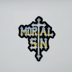 Pode incluir: Patch bordado com a inscrição "MORTAL SIN" em fonte gótica, sobreposta a um desenho de cruz. O patch apresenta uma borda preta e cores amarelo, azul e branco. Ideal para roupas ou acessórios.