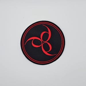 May include: A circular black patch featuring a red triple swirl design within a red-lined border. This fabric patch is designed for application to clothing or accessories, possibly by sewing or ironing.