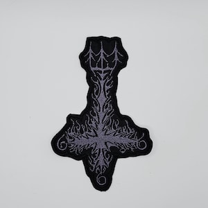 May include: Black patch featuring a stylized inverted cross design with intricate purple embroidery. The patch has a detailed, gothic aesthetic, with a textured appearance. The design includes flame-like elements and decorative swirls.