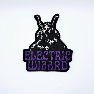 May include: An embroidered patch in black and purple, depicting a horned figure with red eyes. The words "ELECTRIC WIZARD" are in purple block letters. This patch is designed for application on clothing or accessories.