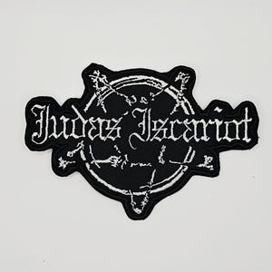 以下が含まれることがあります： ゴシックフォントで「Judas Iscariot」と白く刺繍された黒いワッペン。中央に星のようなシンボルが描かれた円形のエンブレムと、複雑な白いラインワークが特徴で、全体が太い黒のステッチで縁取られています。