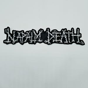 以下が含まれることがあります： 黒い長方形の布製パッチで、白い糸でNAPALM DEATHという文字が刺繍されています。文字はギザギザとしたダメージ加工風のデザインで、黒い背景の上にパッチの幅いっぱいに配置されています。