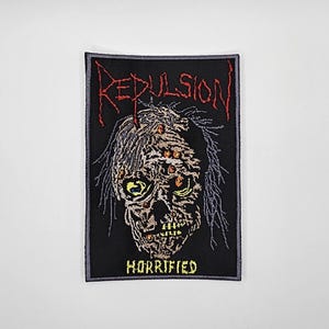 Może przedstawiać: Czarna, haftowana naszywka z motywem czaszki zombie. Słowo "REPULSION" jest wyhaftowane na czerwono nad czaszką, a "HORRIFIED" na żółto pod spodem. Czaszka ma żółte oczy i szare włosy.