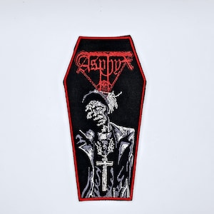 May include: Black coffin-shaped patch with a red border. The patch features a detailed embroidered image of a figure wearing a hat and cross pendant. The word "Asphyx" is embroidered in red at the top.