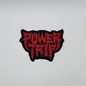 May include: A black and red embroidered patch with the words "POWER TRIP" in a stylized font. The text is in red, with a black border. The patch has a unique, jagged shape.