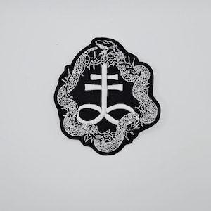 Può includere: Toppa nera con un disegno ricamato bianco. Il disegno presenta una croce con una base a doppia barra, circondata da due serpenti. La toppa ha un bordo nero ed è posta su uno sfondo bianco.