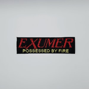May include: Black rectangular patch featuring the word "EXUMER" in red, distressed lettering. Beneath, in yellow, is the phrase "POSSESSED BY FIRE". The patch is designed to be sewn onto clothing or bags.